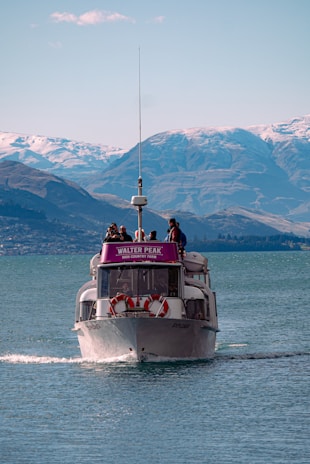 A boat named Walter Peak High Country Farm navigates a serene body of water, with a backdrop of majestic, snow-capped mountains under a clear blue sky. Several people are visible on the deck of the boat, enjoying the scenic view.