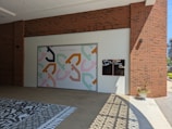 A large mural with an abstract geometric pattern in pastel colors decorates a wall. Above the mural, the phrase 'Kindness is Contagious' is written. Nearby, there are two small chalkboard panels with text that reads 'Take what you Need' and 'Give what you Can'. The building features red brick walls and the ground has patterned tiles. A small plant in a white pot is positioned in sunlight, which casts intricate shadows on the ground.