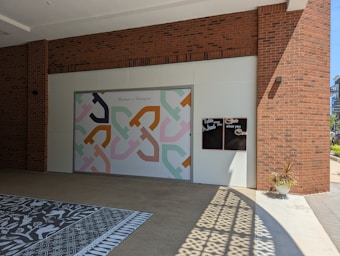 A large mural with an abstract geometric pattern in pastel colors decorates a wall. Above the mural, the phrase 'Kindness is Contagious' is written. Nearby, there are two small chalkboard panels with text that reads 'Take what you Need' and 'Give what you Can'. The building features red brick walls and the ground has patterned tiles. A small plant in a white pot is positioned in sunlight, which casts intricate shadows on the ground.