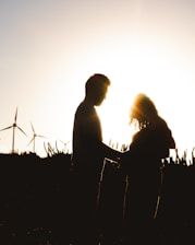 A silhouetted couple faces each other with the sun setting behind them, creating a warm glow. Wind turbines are visible in the background, adding a sense of open landscape and environmental awareness.