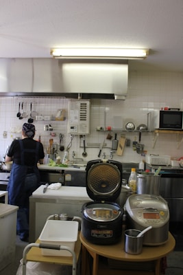 A kitchen with a person standing at the sink wearing a dark shirt and apron, possibly washing or preparing something. The countertop is organized with various utensils hanging on the wall. There are two rice cookers on a small table in the foreground. The room has fluorescent lighting and white tiled walls, with a microwave and other cooking equipment visible.