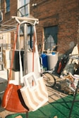 Two handbags hang on a metal stand in an outdoor setting. Behind them, there is an assortment of objects including a bicycle, containers, and various items against a brick wall of a building. There are shadows and green artificial grass on the ground.