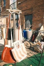 Two handbags hang on a metal stand in an outdoor setting. Behind them, there is an assortment of objects including a bicycle, containers, and various items against a brick wall of a building. There are shadows and green artificial grass on the ground.