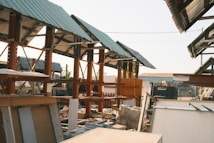 A construction site with several wooden structures under green metal roofs. There are scattered boards and construction materials around. The setting has a slightly industrial feel, with a mix of completed and incomplete elements.
