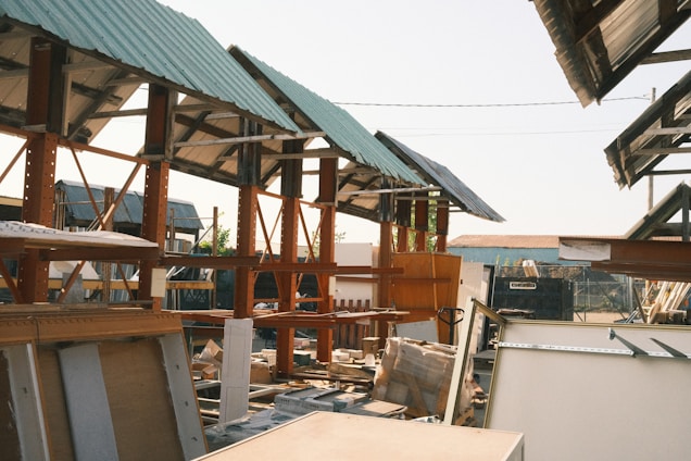 A construction site with several wooden structures under green metal roofs. There are scattered boards and construction materials around. The setting has a slightly industrial feel, with a mix of completed and incomplete elements.