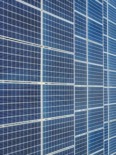 A large array of solar panels with a grid pattern, featuring dark blue and reflective surfaces aligned in a structured manner. The panels are arranged side by side, creating a uniform and organized layout.