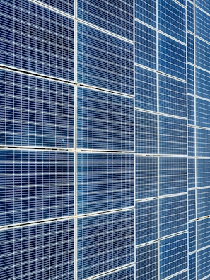A large array of solar panels with a grid pattern, featuring dark blue and reflective surfaces aligned in a structured manner. The panels are arranged side by side, creating a uniform and organized layout.