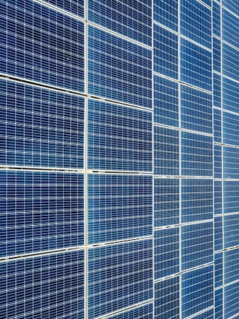 A large array of solar panels with a grid pattern, featuring dark blue and reflective surfaces aligned in a structured manner. The panels are arranged side by side, creating a uniform and organized layout.