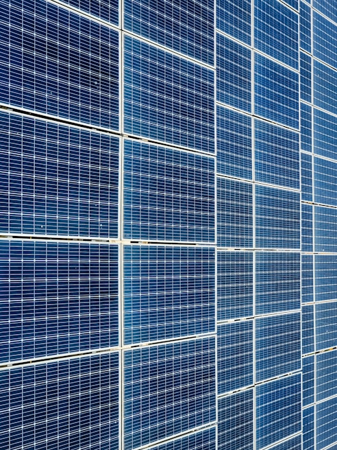 A large array of solar panels with a grid pattern, featuring dark blue and reflective surfaces aligned in a structured manner. The panels are arranged side by side, creating a uniform and organized layout.