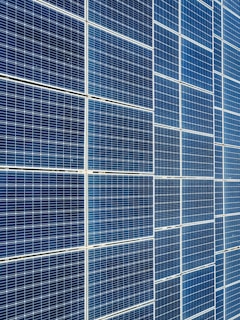 A large array of solar panels with a grid pattern, featuring dark blue and reflective surfaces aligned in a structured manner. The panels are arranged side by side, creating a uniform and organized layout.