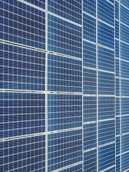 A large array of solar panels with a grid pattern, featuring dark blue and reflective surfaces aligned in a structured manner. The panels are arranged side by side, creating a uniform and organized layout.