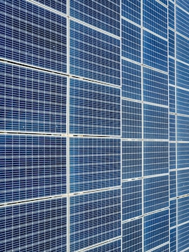 A large array of solar panels with a grid pattern, featuring dark blue and reflective surfaces aligned in a structured manner. The panels are arranged side by side, creating a uniform and organized layout.