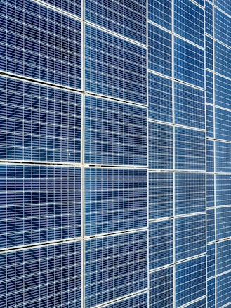 A large array of solar panels with a grid pattern, featuring dark blue and reflective surfaces aligned in a structured manner. The panels are arranged side by side, creating a uniform and organized layout.