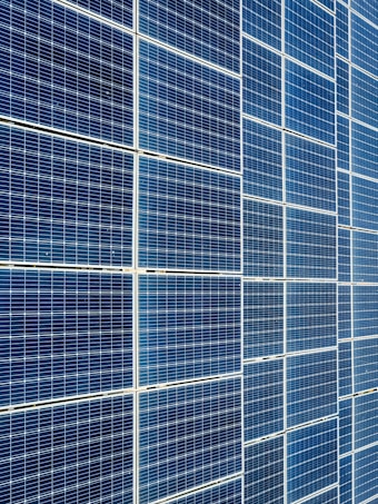 A large array of solar panels with a grid pattern, featuring dark blue and reflective surfaces aligned in a structured manner. The panels are arranged side by side, creating a uniform and organized layout.