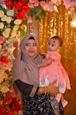 A woman dressed in a traditional outfit and hijab holds a baby wearing a pink dress. They stand in front of a decorative background featuring roses in various colors and glowing lights, creating a festive atmosphere.