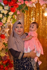 A woman dressed in a traditional outfit and hijab holds a baby wearing a pink dress. They stand in front of a decorative background featuring roses in various colors and glowing lights, creating a festive atmosphere.