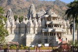 An intricately designed white marble temple with numerous spires and detailed carvings stands against a backdrop of rolling hills. The well-maintained garden and tall palm tree accentuate the temple's grandeur.