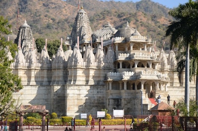 An intricately designed white marble temple with numerous spires and detailed carvings stands against a backdrop of rolling hills. The well-maintained garden and tall palm tree accentuate the temple's grandeur.