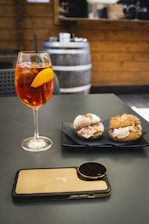 A glass of Aperol Spritz with an orange slice on a table, accompanied by two small sandwiches on a black napkin and a smartphone with a camera attachment. The background features a rustic setting with a wooden barrel and a counter, suggesting a casual dining atmosphere.