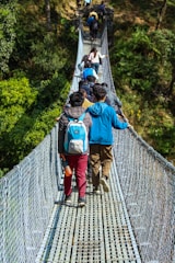 A group of people is walking along a narrow, suspended footbridge surrounded by lush greenery. Many of them carry backpacks, and there is a noticeable sense of movement as they proceed in single file.
