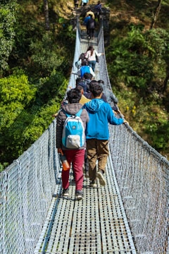A group of people is walking along a narrow, suspended footbridge surrounded by lush greenery. Many of them carry backpacks, and there is a noticeable sense of movement as they proceed in single file.