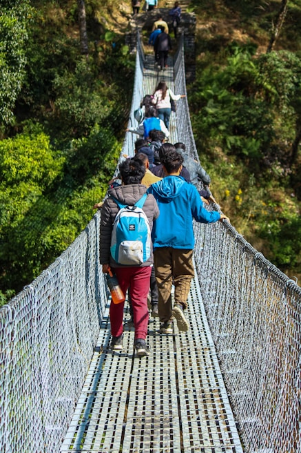 Trekking group crossing a wooden bridge over a rushing mountain stream surrounded by lush greenery.