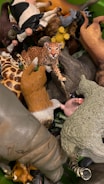 A collection of various animal figurines is piled together, featuring a leopard prominently in the center. Other recognizable animals include a cow, giraffe, and possibly a sheep, among others. The figurines are crafted with distinct colors and details to resemble actual animals.