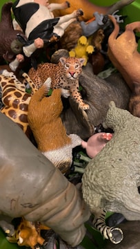 A collection of various animal figurines is piled together, featuring a leopard prominently in the center. Other recognizable animals include a cow, giraffe, and possibly a sheep, among others. The figurines are crafted with distinct colors and details to resemble actual animals.