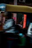 A blurred image capturing the motion of a rickshaw moving past a wall covered with posters and advertisements. The scene is dark with the background details slightly obscured by motion blur, and the rickshaw's canopy is a prominent yellow.