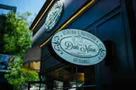 A storefront sign displaying 'Don Nino' with decorative elements and text indicating it is a gelateria and pasticceria, located on a stylish, dark-colored building exterior with trees in the background.
