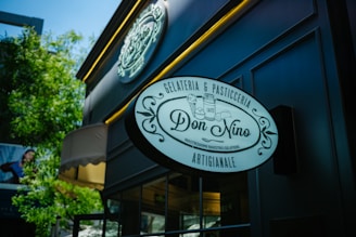 A storefront sign displaying 'Don Nino' with decorative elements and text indicating it is a gelateria and pasticceria, located on a stylish, dark-colored building exterior with trees in the background.