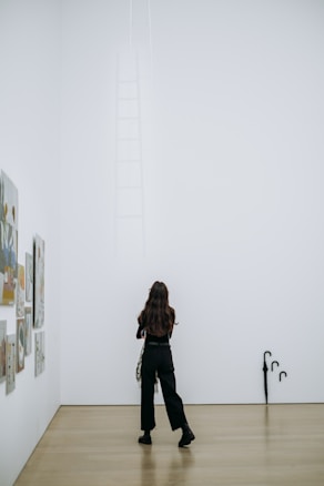 A person with long hair stands in a bright, minimalist gallery space, looking at an installation on the wall. The installation includes a barely visible ladder reaching upwards. Various framed artworks are displayed on the left wall, and there are two black umbrellas leaning against the right wall near the floor.
