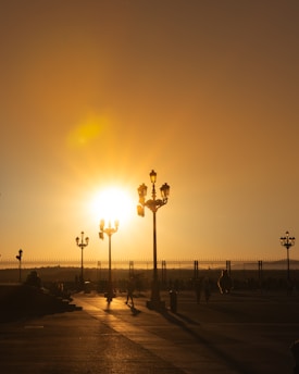 A sunset over a plaza with ornate lampposts casting long shadows on the pavement. The sky is a warm gradient of orange as people walk leisurely, creating a serene silhouette scene.