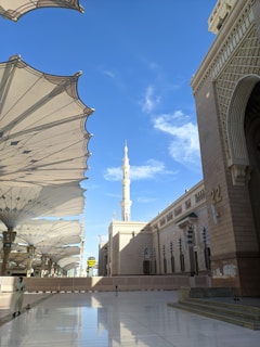 A large mosque with a prominent minaret stands under a clear blue sky. Intricate architectural details adorn the walls, and large, white, umbrella-like structures provide shade in the open courtyard. A person in traditional attire walks near the edge of the courtyard.
