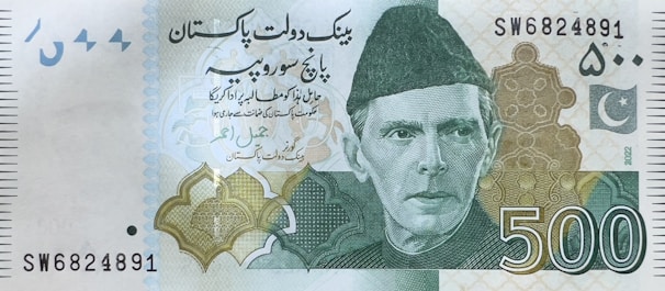 A 500 currency note featuring a portrait of a man wearing a traditional cap. The note includes text in a foreign script, intricate geometric patterns, and a crescent and star emblem. The serial number is visible at the upper right and lower left corners.