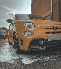 A bright yellow car is parked outdoors, covered in soap suds as if being washed. The image captures a low-angle view focusing mainly on the front of the car, revealing the distinctive details like the headlights and the logo on the grille. The surrounding area appears to be a parking space next to a brick building with other vehicles in the background.