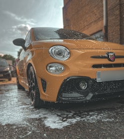 A bright yellow car is parked outdoors, covered in soap suds as if being washed. The image captures a low-angle view focusing mainly on the front of the car, revealing the distinctive details like the headlights and the logo on the grille. The surrounding area appears to be a parking space next to a brick building with other vehicles in the background.