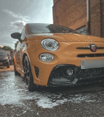 A bright yellow car is parked outdoors, covered in soap suds as if being washed. The image captures a low-angle view focusing mainly on the front of the car, revealing the distinctive details like the headlights and the logo on the grille. The surrounding area appears to be a parking space next to a brick building with other vehicles in the background.