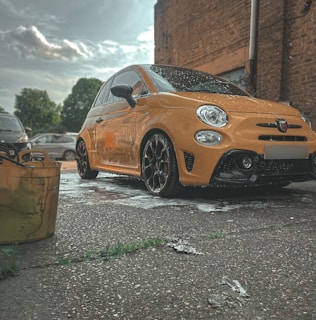 A bright yellow compact car is parked on a wet pavement, likely after a car wash. The car has stylish black alloy wheels and is positioned in an outdoor setting with a brick wall to the right and parked cars in the background. On the left side, a yellow bucket is visible, suggesting cleaning activities.