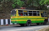 A colorful bus painted in yellow and green is traveling on a road lined by trees and a rocky wall. The bus has signboards indicating 'Birpara' and 'Siliguri,' with passengers visible through the windows, some of whom are standing. The side of the road is bordered by black and white striped barriers.