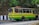 A colorful bus painted in yellow and green is traveling on a road lined by trees and a rocky wall. The bus has signboards indicating 'Birpara' and 'Siliguri,' with passengers visible through the windows, some of whom are standing. The side of the road is bordered by black and white striped barriers.