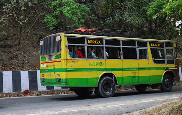 A colorful bus painted in yellow and green is traveling on a road lined by trees and a rocky wall. The bus has signboards indicating 'Birpara' and 'Siliguri,' with passengers visible through the windows, some of whom are standing. The side of the road is bordered by black and white striped barriers.