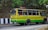 A colorful bus painted in yellow and green is traveling on a road lined by trees and a rocky wall. The bus has signboards indicating 'Birpara' and 'Siliguri,' with passengers visible through the windows, some of whom are standing. The side of the road is bordered by black and white striped barriers.