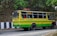 A colorful bus painted in yellow and green is traveling on a road lined by trees and a rocky wall. The bus has signboards indicating 'Birpara' and 'Siliguri,' with passengers visible through the windows, some of whom are standing. The side of the road is bordered by black and white striped barriers.