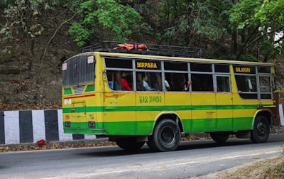 A colorful bus painted in yellow and green is traveling on a road lined by trees and a rocky wall. The bus has signboards indicating 'Birpara' and 'Siliguri,' with passengers visible through the windows, some of whom are standing. The side of the road is bordered by black and white striped barriers.