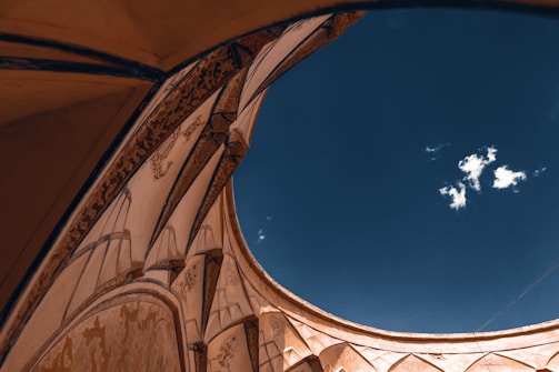 An architectural detail of a building highlights ornate archways and decorative designs against a backdrop of clear blue sky with a few white clouds. The structure suggests elements of historic or classical design.