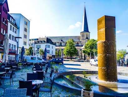 The image features an outdoor urban scene with a cobblestone plaza, outdoor seating, and a water fountain. In the background, there is a historic church with a tall spire. Flanking the square are various buildings, including shops and residential structures, while trees provide greenery.