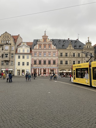 An urban scene with historic buildings featuring intricate architectural details. The area is paved with cobblestones, and several people are walking across the open space. A yellow tram is passing on the right. The sky is overcast, creating a subdued atmosphere.