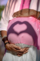A pregnant person is shown with hands forming a heart shape shadow on their pink covered belly, with the person's hands placed underneath the belly. The person is wearing a light pink shirt over a darker pink top and a white skirt. There is a pink ribbon visible on the clothing.