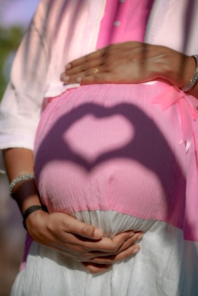 A pregnant person is shown with hands forming a heart shape shadow on their pink covered belly, with the person's hands placed underneath the belly. The person is wearing a light pink shirt over a darker pink top and a white skirt. There is a pink ribbon visible on the clothing.